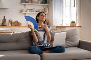 Woman sitting on a couch with a laptop, using a blue fan to cool herself, illustrating the need for effective air conditioning and comfort in home environments.