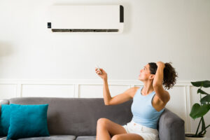 Woman sitting on a couch looking frustrated at a malfunctioning air conditioning unit on the wall, remote control in hand, illustrating common signs of AC issues.