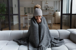 Person wrapped in a gray blanket and wearing a knitted hat, sitting on a couch, appearing cold and uncomfortable in a cozy indoor setting, highlighting the importance of effective heating solutions during winter.